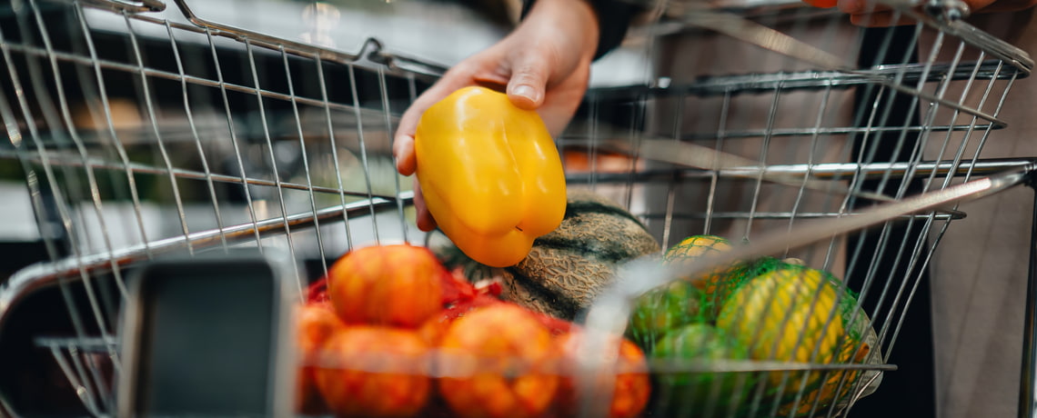 Person legt gelbe Paprika in Einkaufskorb mit Obst.