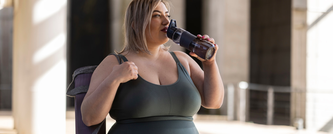 Übergewichtige Frau in Sportkleidung trinkt Wasser aus einer Trinkflasche.